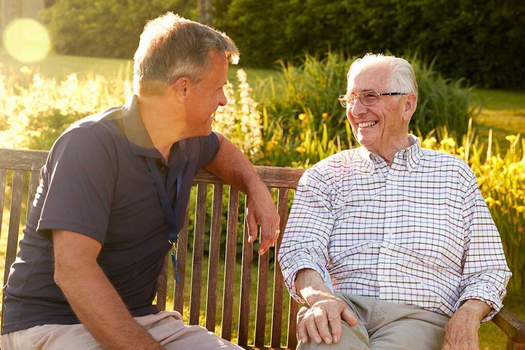 Two older adults looking at a laptop together and discussing printouts about living options in a cozy, sunlit living room.